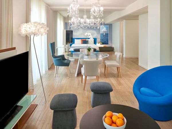 A modern, spacious living area with a blue accent chair, round dining table, white chairs, chandelier, a TV, and a bowl of oranges on a dark table, all in a bright, airy room.