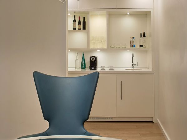 A modern, chic lounge area with a blue accent chair, glass table, and a white kitchenette in the background, under a crystal chandelier.