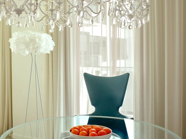 A modern dining area with a glass round table, white chairs, a teal chair, a crystal chandelier, sheer curtains, and a bowl of red apples on the table.
