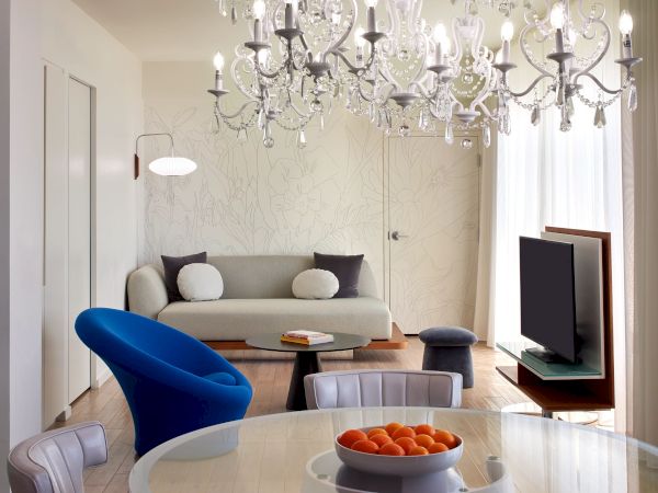 A stylish living room with a large chandelier, a gray sofa, blue accent chair, round dining table with oranges, and a flat-screen TV in a bright, modern space.