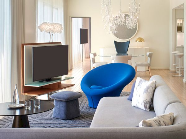 A modern living room with a gray sofa, blue accent chair, coffee table and flat-screen TV, bright natural light, and a chic dining area in the background.