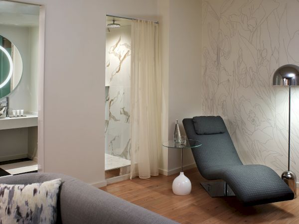 A modern, stylish living area with a gray chaise lounge, patterned wallpaper, a tall arc floor lamp, and a glass-walled shower nook visible in the background.
