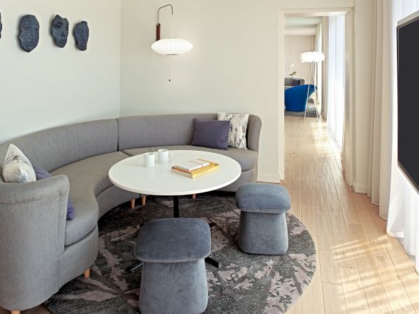 Cozy, modern living room with a curved gray sofa, round white coffee table, small ottomans, geometric wall art, and a bright hallway beyond.