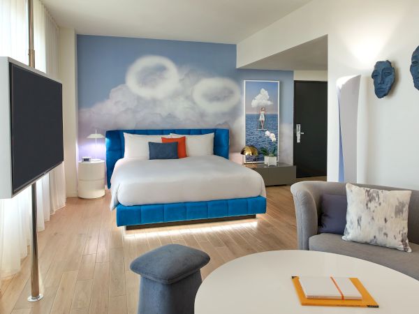 A bright blue-themed hotel room with a king bed, round wall art, sea-blue accents, a gray sofa, round table, and a large TV on the left.