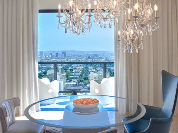 A bright, modern dining nook with a glass round table, white chairs, a blue accent chair, chandeliers, sheer curtains, and a cityscape view from a balcony.