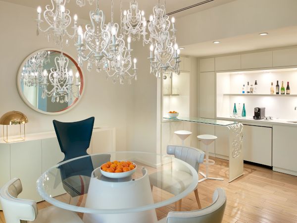 A stylish, bright dining nook with a glass round table and white chairs, chandelier overhead, a bar area in the back, and a round mirror on the wall.