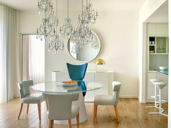 A bright, modern dining room with a glass table and six chairs, chandeliers overhead, and a light, airy feel. The open kitchen is visible in the background.