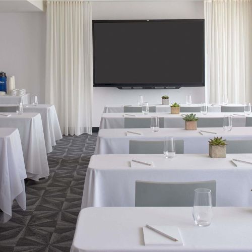 A modern conference room set for a formal event with white-clothed tables, small plants, and a large screen at the front ending the sentence.