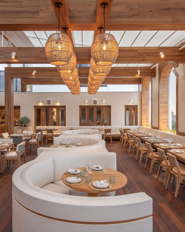 A stylish, airy restaurant with warm wood tones, curved white seating, and long communal tables under woven pendant lights, creating a cozy outdoor‑in vibe.
