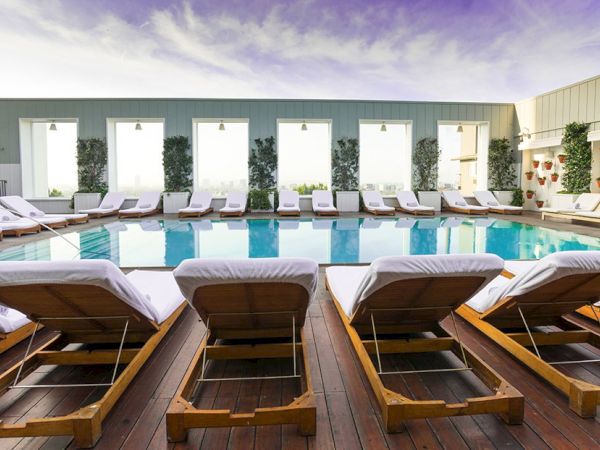 A rooftop hotel pool area with sun loungers facing a rectangular pool, white cushions, and a modern, airy backdrop under a blue sky.