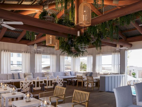 A stylish indoor lounge under wooden beams, with hanging greenery, lantern lights, fans, and cushioned seating.