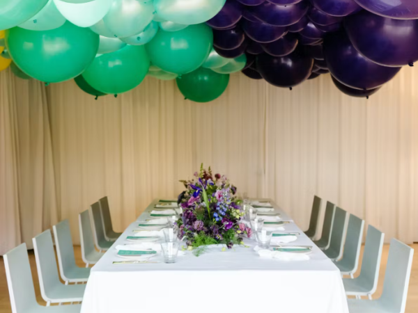 A long dining table with white tablecloth, silverware, chairs, and a purple-green balloon arch overhead, a floral centerpiece in the middle.