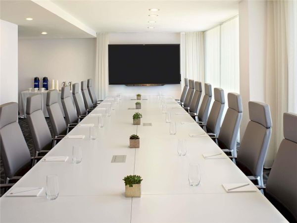 A modern conference room with a long white table, gray ergonomic chairs, water bottles, plants, and a large screen at one end, ready for a meeting.