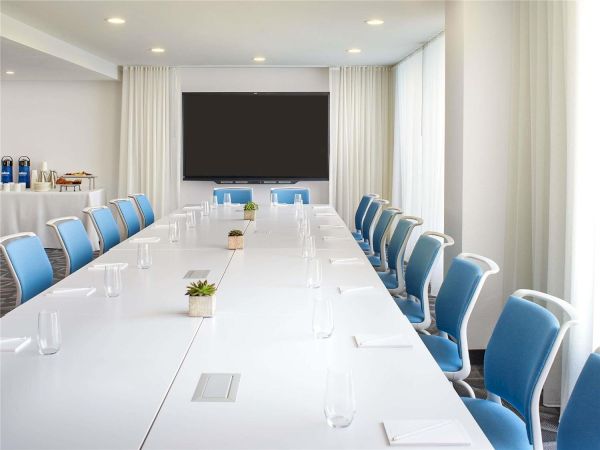 A bright, modern conference room with a long white table, blue chairs, and a large projector screen at the far end, ready for a meeting.