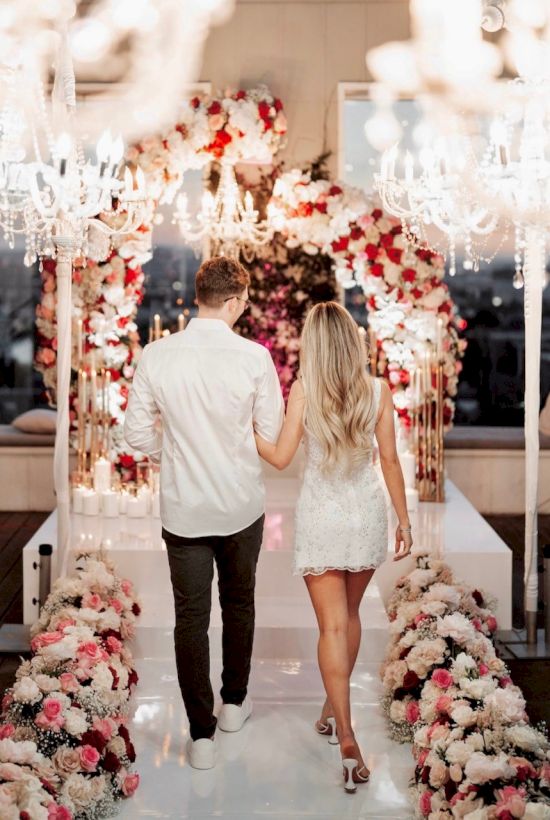 A couple in a white wedding dress and white shirt walk hand-in-hand down a floral, chandelier-lit aisle at a wedding venue, romantic ambiance.