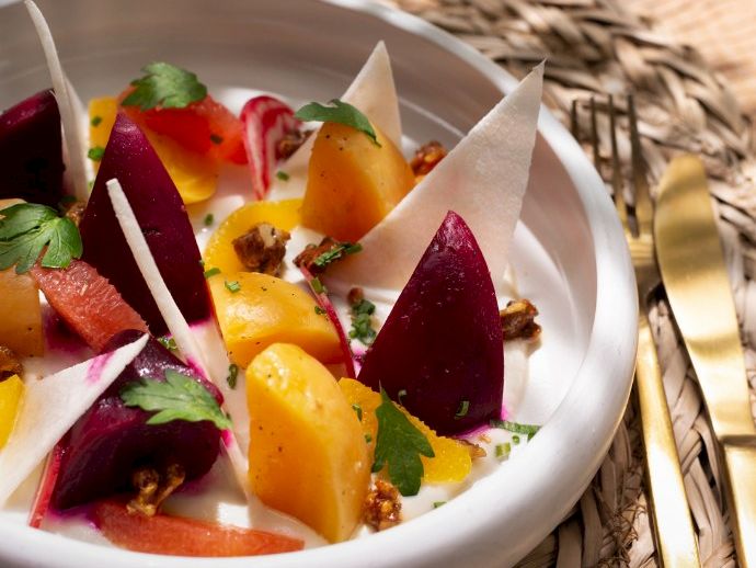 A colorful fruit and white chocolate dessert with beetroot-like pieces, mint leaves, sesame seeds, and almond shards on a white bowl.