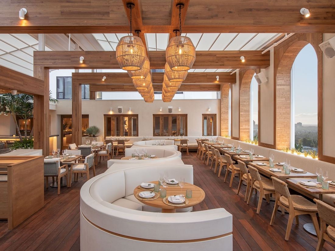 A stylish restaurant with warm wooden beams, round booths, and long communal tables under woven pendant lights, bright and airy interior.