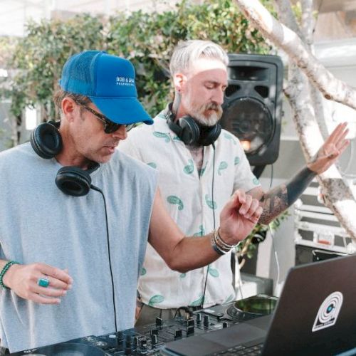 Two DJs perform outdoors, wearing headphones by a laptop and mixer, one gesturing with a raised finger as the other cues the crowd.