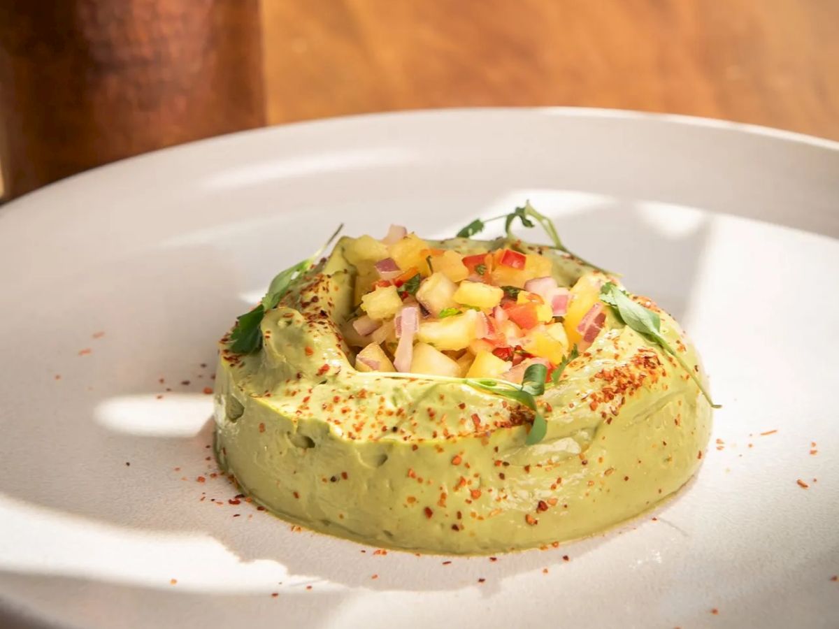 Guacamole topped with diced salsa, served on a white plate with plantain chips in the background, sprinkled with chili flakes.