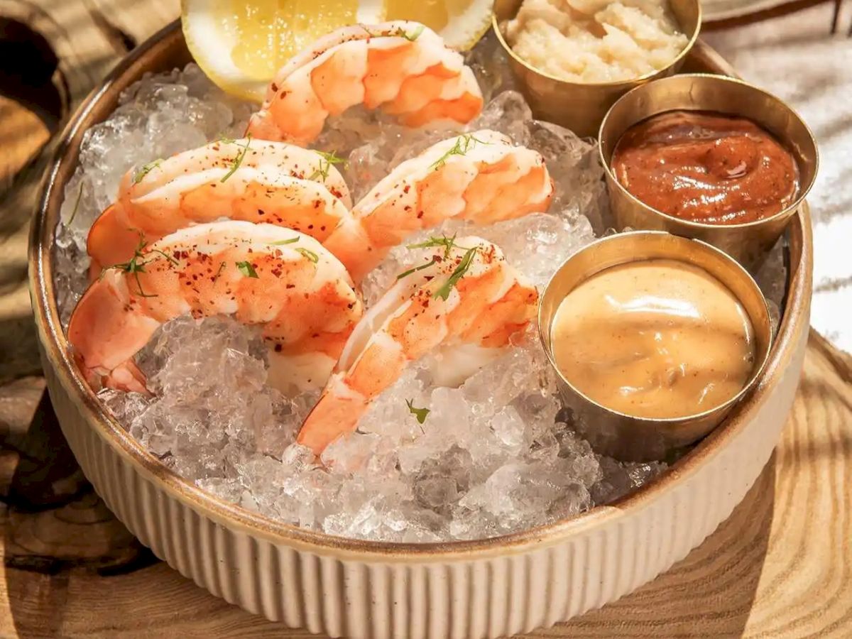 Shrimp cocktail on ice with lemon wedges and dipping sauces on the side; served in a round ribbed bowl over a wooden board.