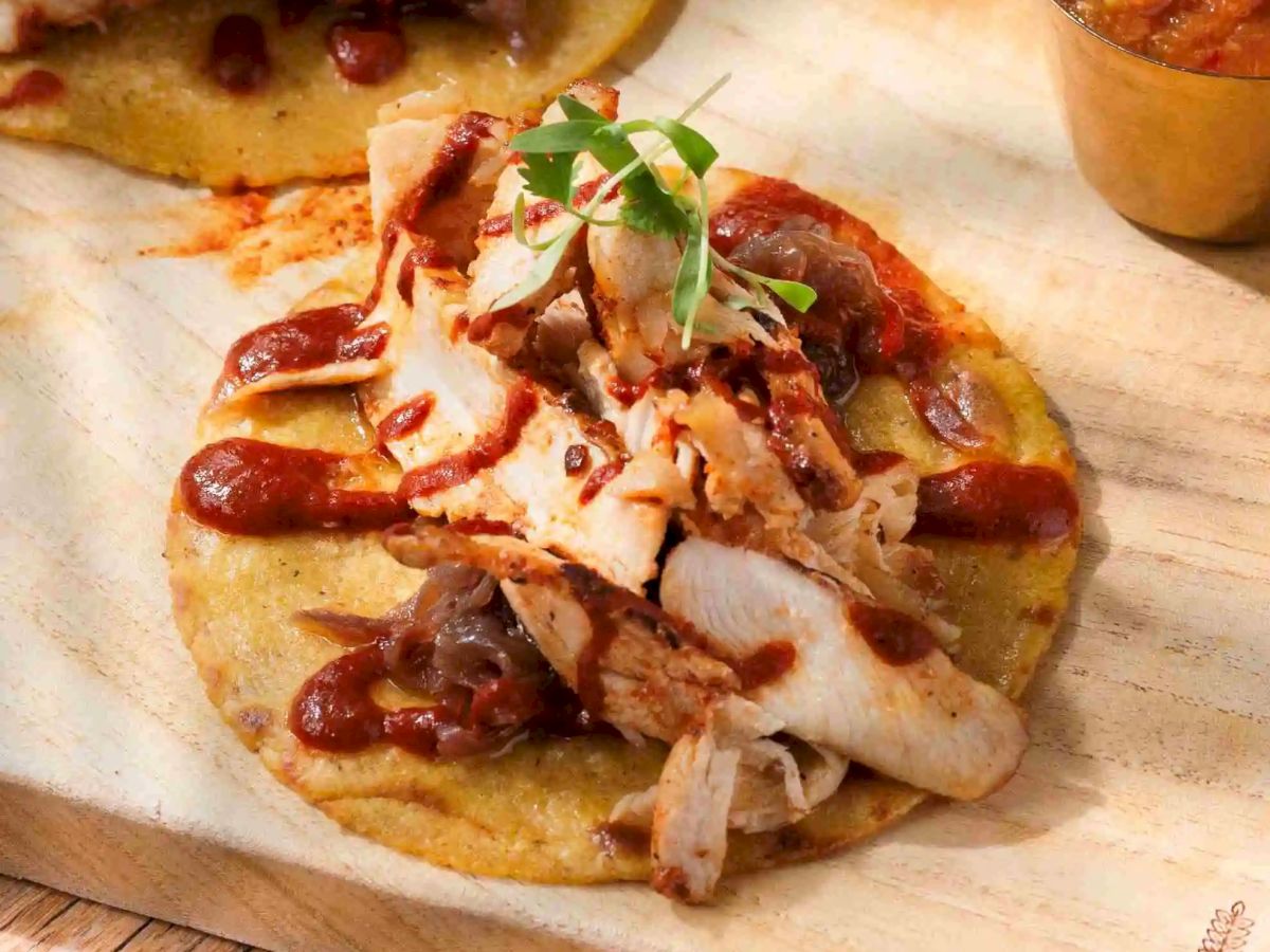 Two short rib or chicken tacos topped with shredded meat, drizzled sauce, and herbs on corn tortillas, served with a small side cup of salsa.