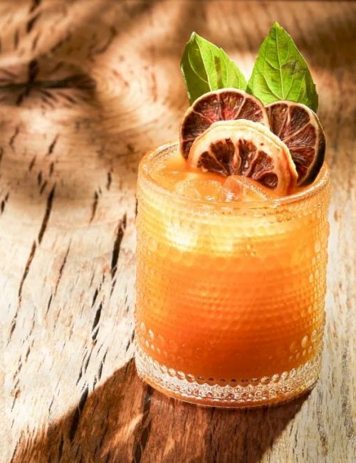 A refreshing orange drink in a glass with lime and dried lemon slices, garnished with mint, on a wooden surface.