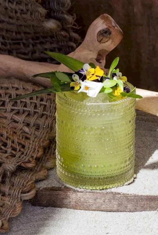 A green cocktail in a textured glass with a flower garnish, rustic wicker and wooden background, sunlit scene, summery vibe.