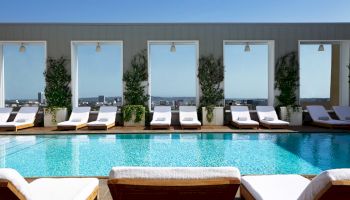 A luxurious rooftop pool scene with sun loungers, wooden deck, potted plants, and large windows reflecting a bright blue sky.