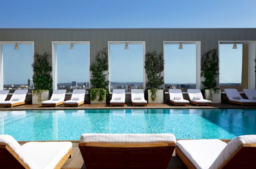 A luxurious rooftop pool scene with sun loungers, wooden deck, potted plants, and large windows reflecting a bright blue sky.
