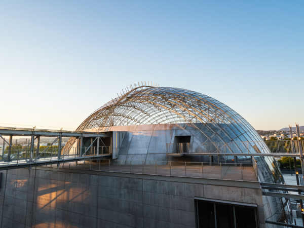 A futuristic glass dome atop a stone building with a curved metal framework, connected by elevated walkways, under a clear blue sky.