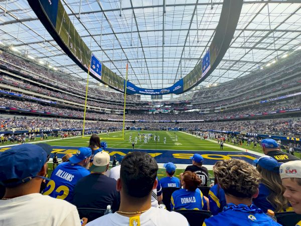 A packed stadium viewed from the stands, players on the field, blue jerseys in the crowd, and a bright overhead roof.
