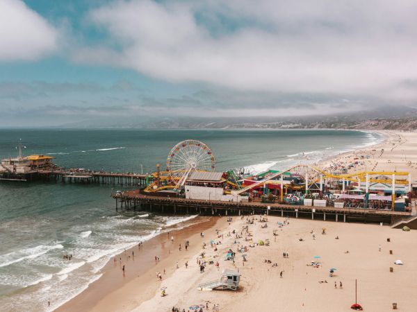 A bustling beach scene with a long pier, colorful carnival rides at a boardwalk, and crowds enjoying sun and waves along the shoreline.