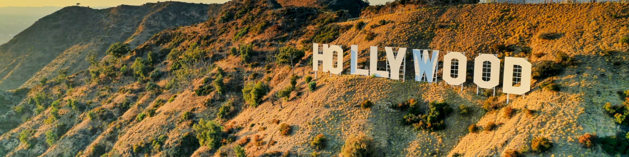 A selfie-friendly hillside with the Hollywood sign perched on rugged terrain under a bright blue sky at golden hour.
