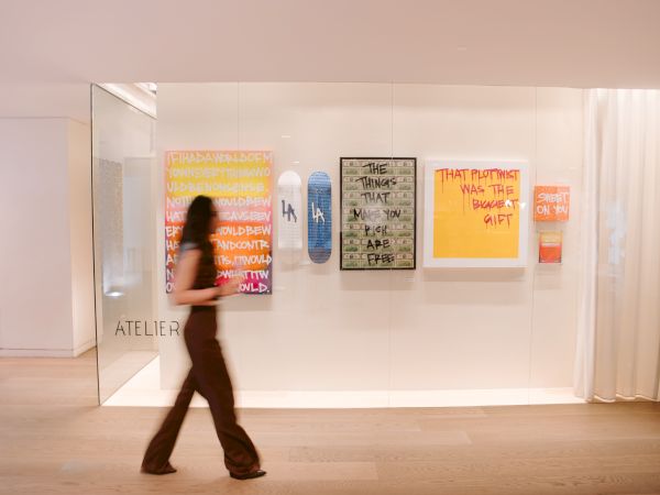 A person walks past a wall with colorful posters in an art gallery, labeled &ldquo;ATELIER&rdquo; on the left, bright abstract text works displayed.