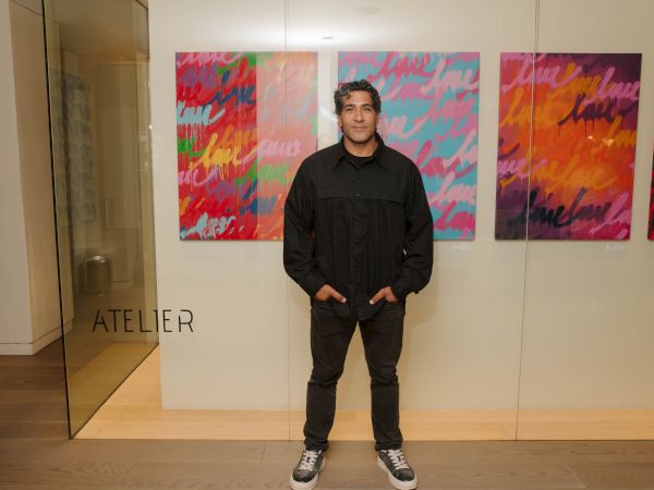 A man stands in an art gallery in front of bright abstract paintings, posing calmly with hands in pockets.