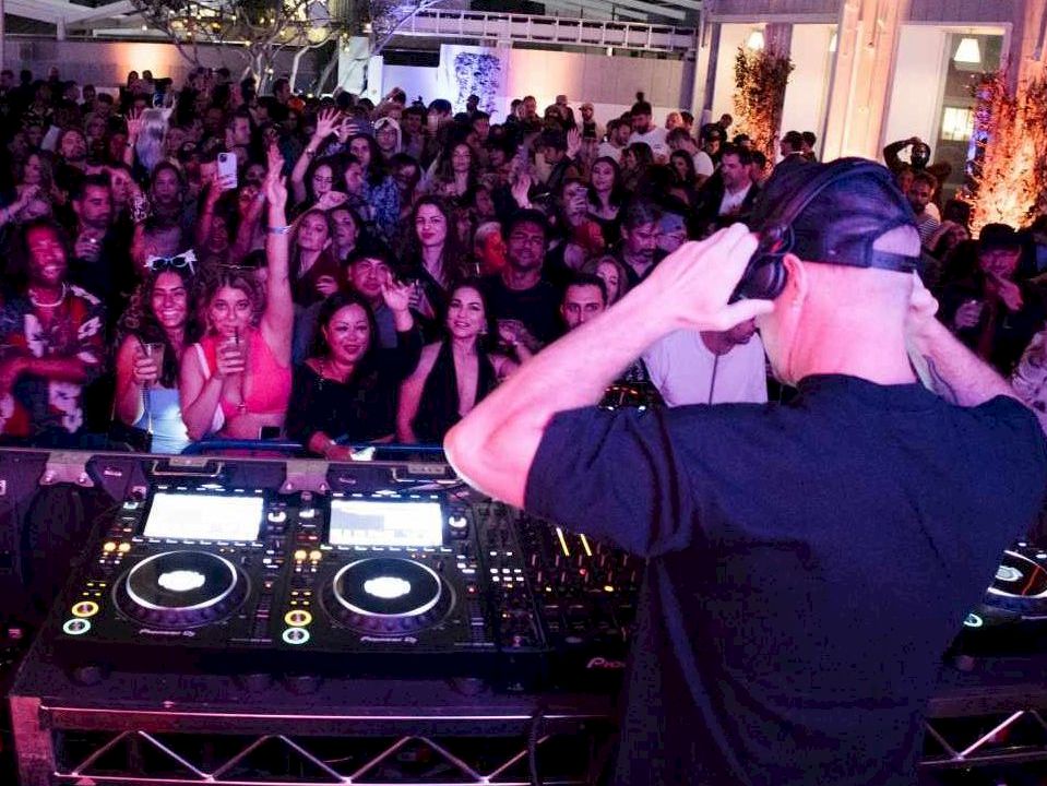 A DJ performs at a crowded indoor party, wearing a cap, blending tracks on turntables while a packed audience cheers and watches the stage.