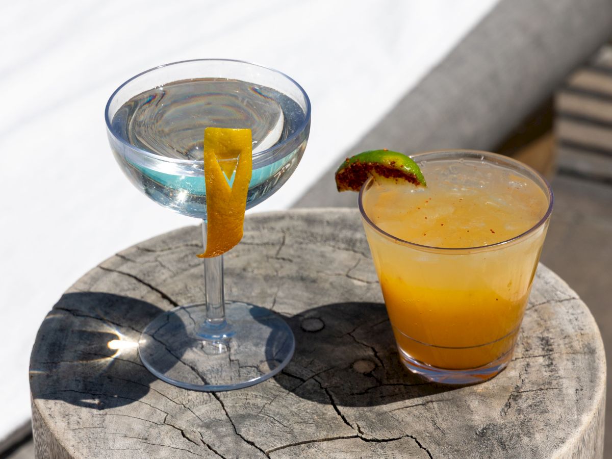 Two cocktails on a weathered wooden stump beside a pool, with blue water, a white towel, and lime/orange garnishes.