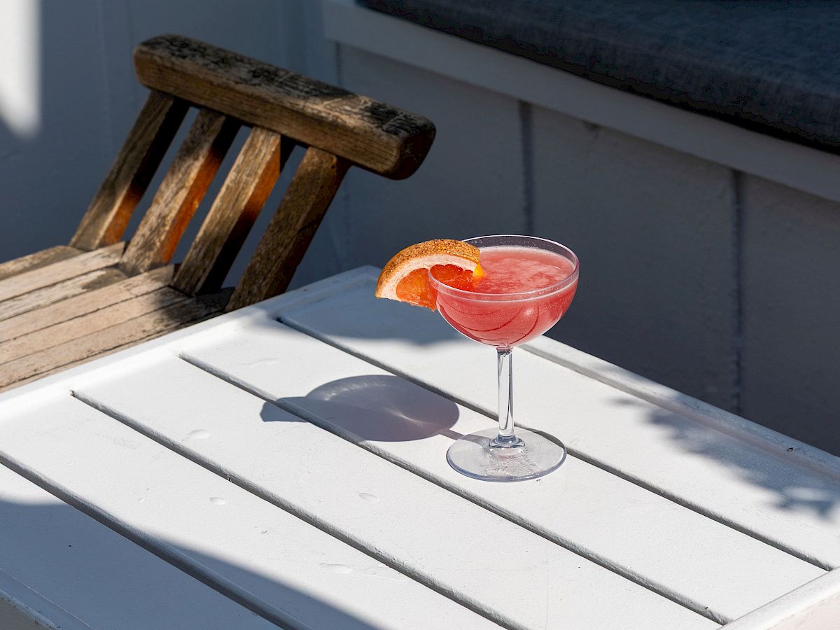 A pink cocktail with an orange slice sits on a white wooden table beside a weathered chair in bright sunlight.