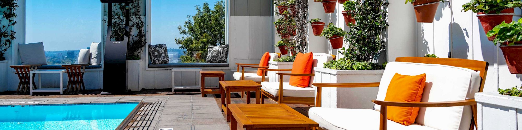 A sunny rooftop patio with a small blue pool, white lounge chairs with orange pillows, and many hanging potted plants.