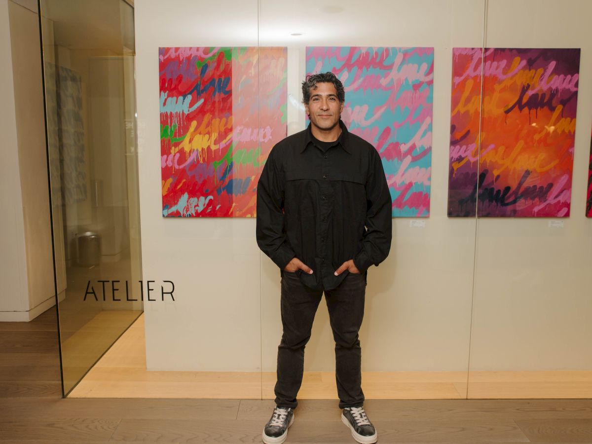 A man stands in an art gallery, posing before colorful abstract paintings, with &ldquo;ATELIER&rdquo; visible by the entrance.