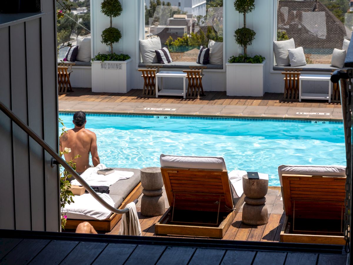 A rooftop pool with lounge chairs, a man in shorts by the pool, white planters, and seating areas with city views in the background.
