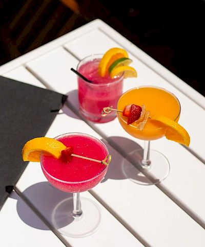 Three bright cocktails with orange wedges and a cherry garnish sit on a white table, a black menu nearby.