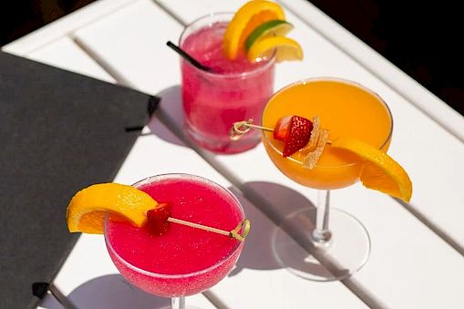Three bright cocktails with orange wedges and a cherry garnish sit on a white table, a black menu nearby.