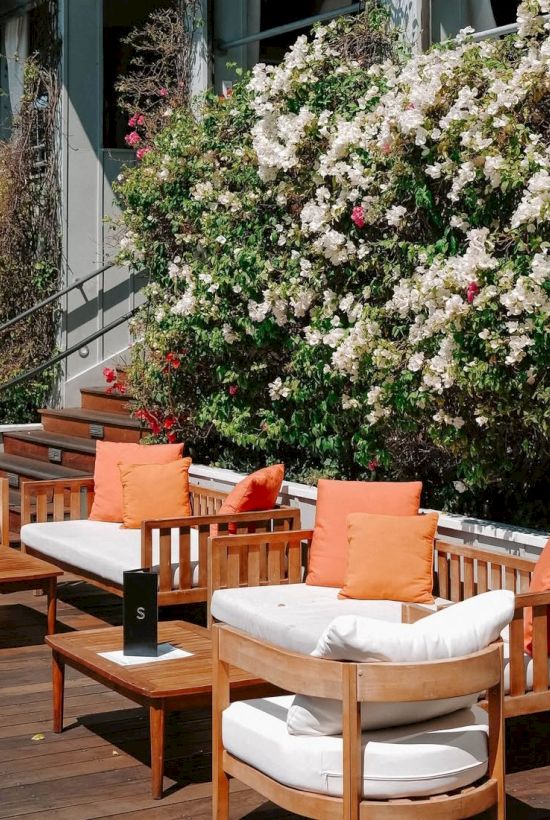 An outdoor patio with wooden furniture and white cushions, orange pillows, and blooming white roses along a railing.