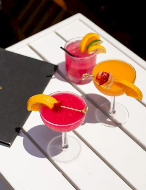 Three cocktail glasses with pink drinks and orange garnish sit on a white table next to a black menu card.