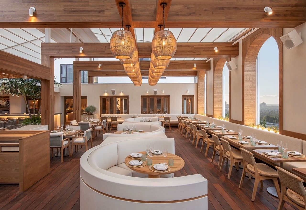 A stylish, bright restaurant with warm wood tones, curved white booth seating, long rows of tables, hanging woven light fixtures, and large windows.