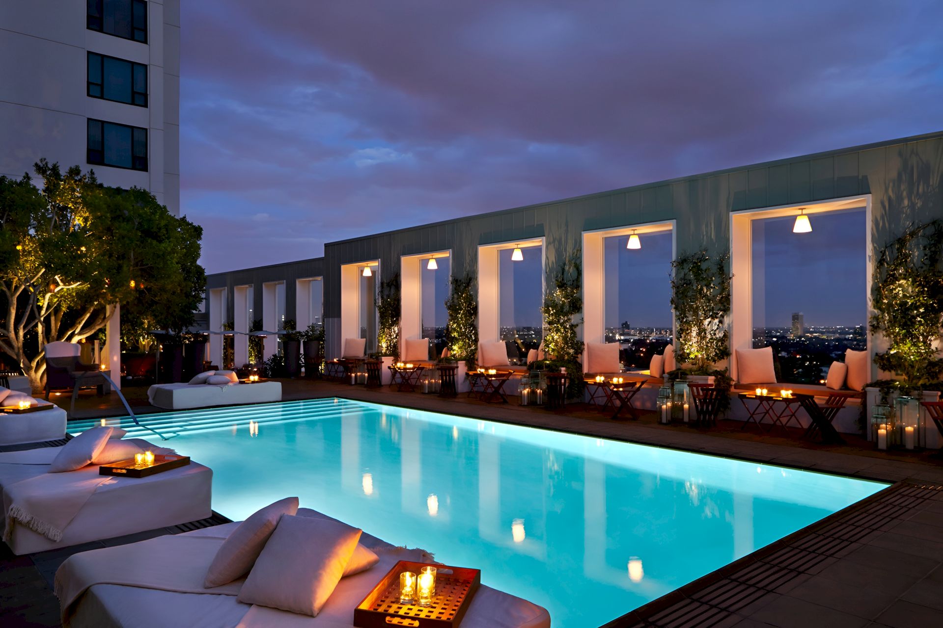 A luxurious rooftop pool area at dusk with lounging chairs, soft lighting, and palm trees, set against a modern building and a purple, cloudy sky.