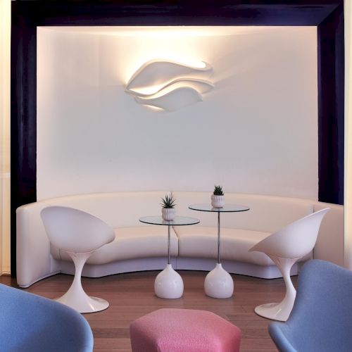 A sleek, modern lounge with a curved white sofa, two white chairs, glass tables, and a soft-lit backdrop featuring a minimalist wave logo.