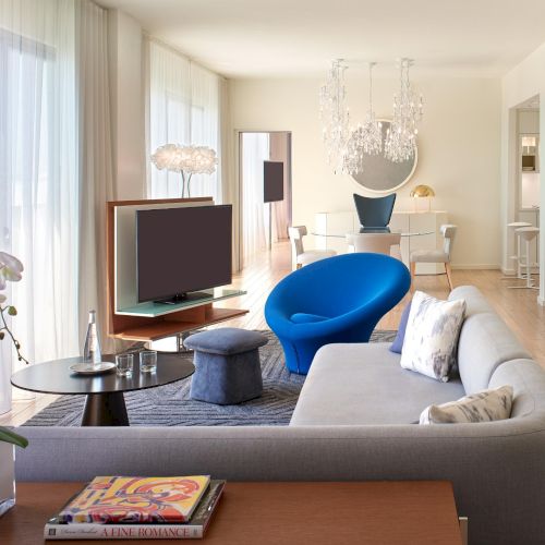 A modern living room with a gray sectional sofa, blue accent chair, glass coffee table, flat-screen TV, potted orchid, and light wood floors.