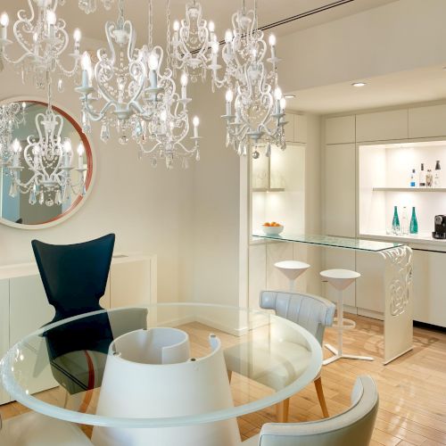 A stylish, bright dining nook with a glass round table, white chairs, crystal chandeliers, and a sleek white kitchenette/bar in the background.